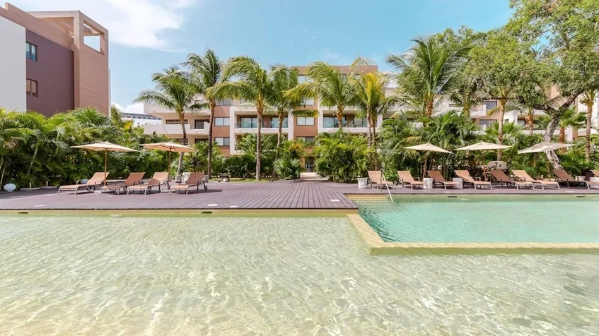 una fachada de un edificio con departamentos, una alberca grande y camastros en lorena ochoa residences playa del carmen